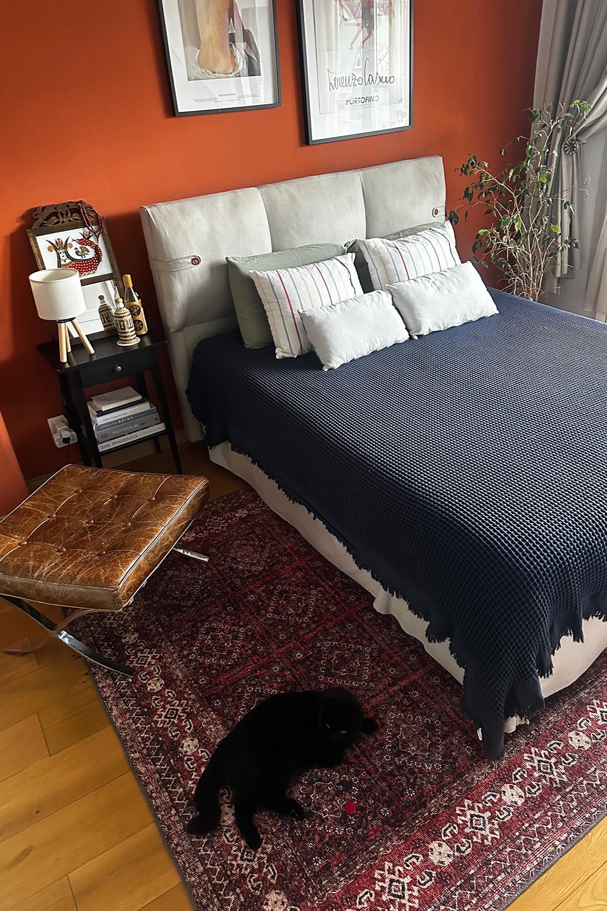 Koyu bordo tonlarında vintage desenli yatak odası halısı, sıcak duvar rengi ve yatak altına yerleşimiyle mekana karakter katan güçlü bir görünüm sunuyor; üzerinde yatan siyah kedi ile birlikte doğal bir atmosfer oluşturuyor